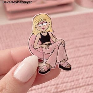 May include: A die-cut sticker featuring a cartoon illustration of a blonde person with glasses, wearing a black tank top and light pink pants, sitting in a pink rounded chair. The person wears pink platform sandals and has a choker necklace.