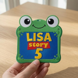 Könnte beinhalten: Ein grüner, froschförmiger Stickaufnäher mit einem blauen rechteckigen Zentrum, das den gelben Text LISA, das Wort story in weißen Buchstaben auf rotem Grund und eine große gelbe Zahl 5 darunter zeigt. Der Frosch hat weiße Augen mit schwarzen Pupillen.
