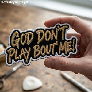 May include: An embroidered patch with a black background and gold lettering that reads GOD DON'T PLAY BOUT ME! The patch features a thick white border and a die-cut shape that follows the contour of the text, creating a bold and readable design.