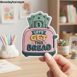 May include: An embroidered patch shaped like a pink toaster with green dollar bills sticking out of the top. The text reads LET'S GET THIS BREAD in yellow and dark green letters, accented with two small pink hearts on a white background border.
