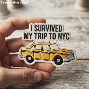 Puede incluir: Un parche bordado que muestra un taxi amarillo con una franja a cuadros blancos y negros en el lateral. Sobre el coche, el texto I SURVIVED MY TRIP TO NYC aparece en letras mayúsculas negras sobre un fondo de color crema.