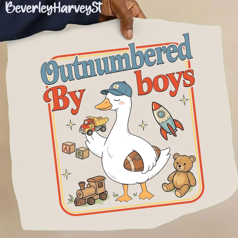 May include: A cream t-shirt with a cartoon goose wearing a cap and holding a toy truck and football. The text "Outnumbered by boys" is above the goose, with other toy graphics.