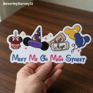Puede incluir: Parche bordado con borde blanco que presenta un sombrero de mago morado, orejas de Mickey Mouse y globos. El parche incluye el texto "Meet Me On Main Street" en azul y rosa, junto con boletos "Magic" y una manzana caramelizada.