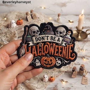 Può includere: Una toppa ricamata nera con il testo DON'T BE A HALLOWEENIE in lettere arancioni. Il design include due teschi, un cappello da strega, una scopa, una zucca, un fantasma e una boccetta di pozione viola con la parola POISON sull'etichetta.