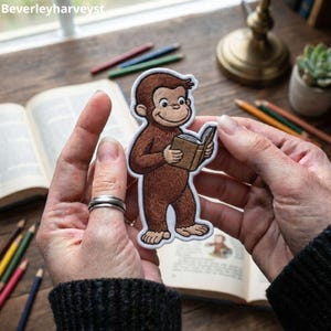 May include: An embroidered patch featuring a brown monkey holding an open tan book. The monkey has a light brown face, ears, hands, and feet, with a white border outlining the entire figure. The design is rendered in detailed thread work.