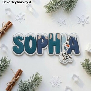 May include: A custom name patch spelling SOPHIA in bold, dark teal and blue letters with white snow-capped tops. The letter I is replaced by a white, cartoon snowman character with a carrot nose, three black buttons, and twig arms.