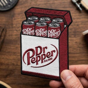 以下が含まれることがあります： Dr Pepperのソーダ缶の箱が描かれた刺繍パッチ。パッチは主に赤と白で、Dr Pepperのロゴが大きく表示されています。「Est.1885」の文字も含まれています。パッチは手で持たれています。