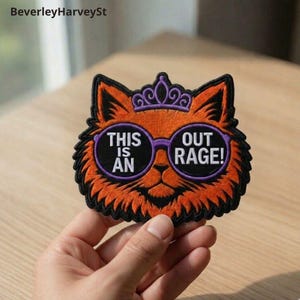 May include: Embroidered patch of an orange cat wearing purple glasses and a crown. The glasses have the words "THIS IS AN OUT RAGE!" in white. The patch has a black border.