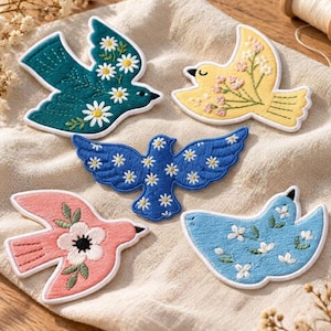 May include: Five embroidered bird patches featuring floral designs. One teal bird with white daisies, one yellow bird with pink flowers, one blue bird with white daisies, one pink bird with a large white flower, and one light blue bird with white blossoms.
