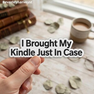 May include: A white die-cut sticker with a black embroidered-style border and black text that reads I Brought My Kindle Just In Case. The sticker has a textured surface and is held against a blurred background of books and a coffee mug.