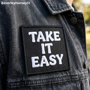 Op de afbeelding: Een zwart vierkant embleem met de woorden "TAKE IT EASY" in het wit geborduurd, bevestigd op een donker spijkerjasje. De patch is gecentreerd op de borst van de jas en biedt een casual en ontspannen uitstraling.