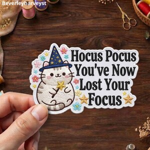 May include: An embroidered patch featuring a light brown tabby cat wearing a blue wizard hat with yellow stars, holding a yellow star wand. The text reads Hocus Pocus You've Now Lost Your Focus, surrounded by small colourful embroidered flowers and stars.