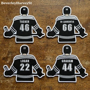 Könnte beinhalten: Vier Hockey-Trikot-Patches in Schwarz, Weiß und Silber. Jeder Patch zeigt den Namen und die Nummer eines Spielers: Tucker 46, Di Laurentis 66, Logan 22 und Graham 44. Die Patches befinden sich auf einer Holzoberfläche.