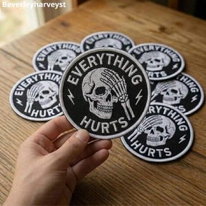 May include: A circular black embroidered patch featuring a white skull with a skeletal hand covering its eyes. The text "EVERYTHING HURTS" is stitched in white capital letters around the skull, accompanied by two small white lightning bolt icons.