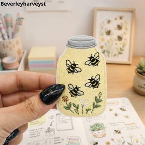 May include: An embroidered patch shaped like a mason jar with a light yellow background, featuring four black and yellow bees, small green stems, and two tiny flowers at the bottom. The jar has a grey lid and a white border edge.