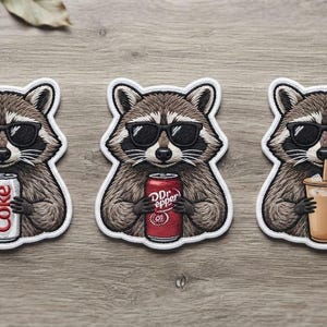 May include: Three embroidered patches featuring raccoons wearing black sunglasses. The first raccoon holds a silver Diet Coke can, the second holds a red Dr Pepper can, and the third holds a tan iced coffee cup with a straw. Each patch has a white border.