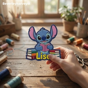 May include: A blue embroidered patch featuring a cartoon character with large ears holding an open pink book, sitting atop a stack of colourful books with the name Lisa printed in yellow letters at the bottom of the design.