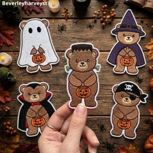 Könnte beinhalten: Fünf bestickte Bärenaufnäher in Halloween-Kostümen: ein Geist, ein Frankenstein-Monster, eine Hexe, ein Vampir und ein Pirat. Jeder braune Bär hält einen kleinen orangefarbenen Kürbis. Die Aufnäher sind auf einer Holzoberfläche angeordnet.