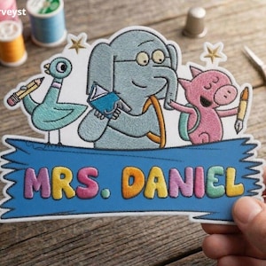 May include: An embroidered patch featuring a blue elephant, a pink pig, and a teal bird with school supplies. The patch includes a blue banner with the text MRS. DANIEL in colourful, raised lettering. The design is detailed with gold stars.