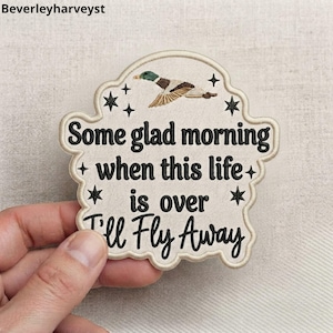 Könnte beinhalten: Ein cremefarbener, bestickter Aufnäher mit gewelltem Rand, der oben eine fliegende Stockente und kleine schwarze Sterne zeigt. Der Text lautet in schwarzer Stickerei: Some glad morning when this life is over I'll Fly Away.