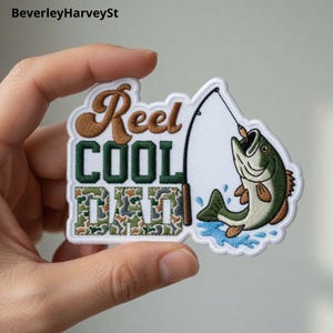 Puede incluir: Parche bordado con las palabras "Reel COOL DAD" en marrón, verde y camuflaje. El parche presenta una caña de pescar y un pez verde con un señuelo en la boca, con detalles de agua azul.