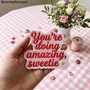 Peut inclure: Un écusson brodé rose clair avec un bord festonné présentant le texte rouge You're doing amazing, sweetie et une petite fleur de marguerite blanche avec un centre jaune dans le coin inférieur droit du motif de l'écusson.