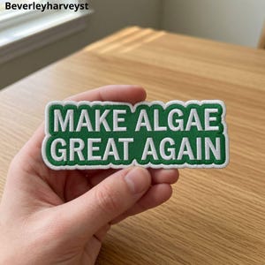 May include: A rectangular embroidered patch with a green background and a white border. The text "MAKE ALGAE GREAT AGAIN" is stitched in white capital letters across the centre of the patch. The edges of the patch follow the shape of the text.