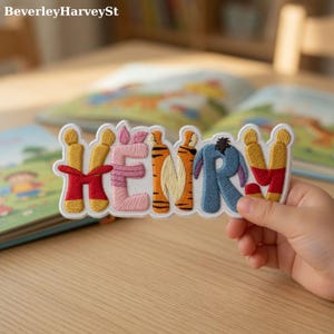 May include: Embroidered patch spelling "HENRY" with letters designed to resemble characters from a children's book. The patch features a white border and is held in front of open books on a wooden surface.