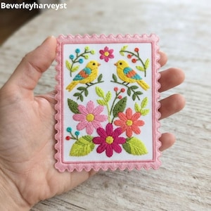 Könnte beinhalten: Ein rechteckiger Stickaufnäher mit einem gewellten rosa Rand auf weißem Stoffgrund. Das Design zeigt zwei gelbe Vögel mit blauen und orangefarbenen Flügeln auf grünen Zweigen, umgeben von rosa, korallenfarbenen und magentafarbenen Blumen und Blättern.