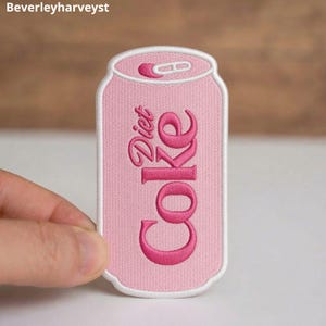 May include: A light pink embroidered patch shaped like a soda can with a white border. The text Diet Coke is stitched in dark pink thread across the center of the patch, with a small tab detail at the top of the can design in matching pink.