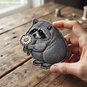 May include: An embroidered patch featuring a grey raccoon with a black eye mask holding a white daisy with a yellow centre. The raccoon has detailed fur texture and is depicted in a seated position. The patch has a dark grey stitched border.