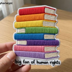 May include: An embroidered patch featuring a stack of seven books in rainbow colors including red, orange, yellow, green, blue, purple, and pink. The white pages are visible on the right side. The text "big fan of human rights" is at the bottom.