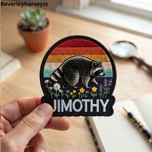 May include: A circular embroidered patch featuring a raccoon walking through a field of small flowers against a sunset background of red, orange, yellow, and teal stripes. The name JIMOTHY is stitched in white capital letters at the bottom.