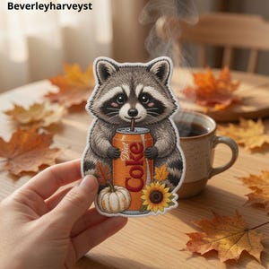 May include: A die-cut sticker featuring a grey and black raccoon holding an orange Diet Coke can with a straw. The raccoon is surrounded by a white pumpkin and a yellow sunflower. The design includes autumn leaves and a white border.
