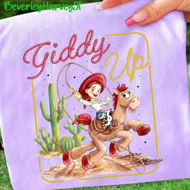 May include: A lilac t-shirt with a cartoon cowgirl riding a horse, and the words "Giddy Up" in red and yellow. The design includes cacti and a brown and white saddle.