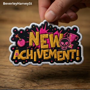 Peut inclure: Écusson brodé avec les mots "NEW ACHIEVEMENT!" en or et rose. L'écusson présente un fond noir avec un crâne, un trophée et un graphique de bombe. L'écusson est tenu par une main.