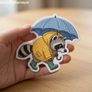 May include: An embroidered patch featuring a raccoon wearing a yellow raincoat and green rain boots while holding a blue umbrella. The design is detailed with textured stitching, showcasing the raccoon's striped tail and masked face in grey and black.