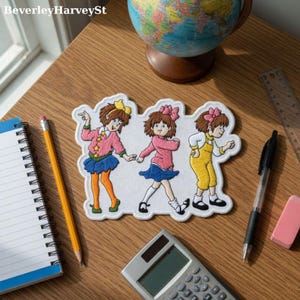 May include: Embroidered patch depicting three cartoon girls in pink, blue, and yellow outfits. The patch has a white border. The image is on a wooden desk with a notebook, pencil, and calculator. A globe is also visible.
