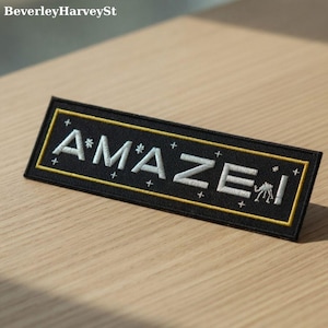 以下が含まれることがあります： 黒い長方形のパッチ。白い文字で「AMAZE！」と刺繍され、黄色の縁取りがあります。パッチの最後には小さな生き物のイラストがあります。パッチは木製の表面にあります。