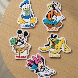 Könnte beinhalten: Fünf gestickte Aufnäher mit Disney-Figuren: Donald Duck beim Lesen, Goofy beim Schreiben, Mickey Maus an einem Schreibtisch, Pluto im Liegen und Minnie Maus beim Lesen. Jeder Aufnäher zeigt den Namen der Figur in bunten Druckbuchstaben.