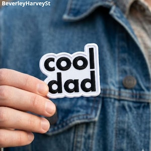 May include: A white embroidered patch with a black border and the black text "cool dad" in a rounded, sans-serif font. The patch features a scalloped edge design that follows the contour of the letters, creating a distinct, readable graphic element.