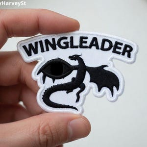 Pode incluir: Patch bordado com a palavra "WINGLEADER" em preto sobre uma faixa branca. O patch apresenta um dragão preto com um olho e asas. O patch tem uma borda branca.