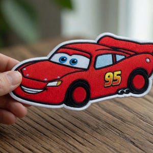 May include: A red embroidered fabric patch shaped like a race car with blue eyes on the windshield, a yellow number 95 on the side, and a white border. The design features black tires, a rear spoiler, and a smiling mouth on the front bumper.