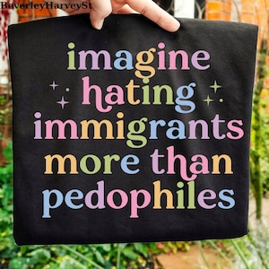 Puede incluir: Camiseta negra con la frase "imagine hating immigrants more than pedophiles" en letras coloridas y estilizadas. El texto está dispuesto en formato apilado. La camiseta está sostenida por una mano.