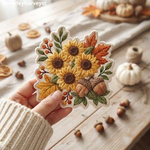 Op de afbeelding: Een geborduurde patch met een groep van drie gele zonnebloemen met bruine harten, twee bruine eikels, oranje bessen en diverse groene en oranje herfstbladeren op een crèmekleurige stoffen achtergrond met een geschulpte rand.