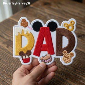 以下が含まれることがあります： 赤と茶色で「DAD」の文字が刺繍されたパッチ。黄色と白の「D」があります。パッチにはミッキーマウスの耳と食べ物をテーマにした装飾が施されています。パッチは手で持たれています。