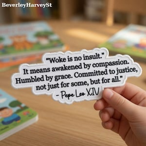 Op de afbeelding: Een witte patch met zwarte geborduurde tekst. De tekst luidt: "Woke is no insult. It means awakened by compassion. Humbled by grace. Committed to justice, not just for some, but for all." - Pepe Leo XIV.