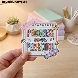 May include: A square embroidered patch featuring a white background with blue lined paper details. The text reads PROGRESS over PERFECTION in colourful letters. It is decorated with a small smiley face, a flower, stars, and two strips of patterned tape.