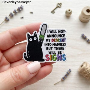 May include: An embroidered patch featuring a black cat holding a gray chainsaw. The white background includes the black text: I WILL NOT ANNOUNCE MY DESCENT INTO MADNESS BUT THERE WILL BE SIGNS, with the words DESCENT and SIGNS in rainbow colors.