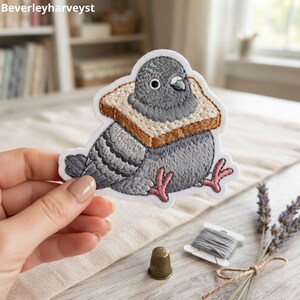 May include: An embroidered patch featuring a grey pigeon with a slice of toasted bread around its neck. The pigeon has dark grey feathers, a white eye, and pink feet. The patch has a white border and is shaped to follow the outline of the bird.